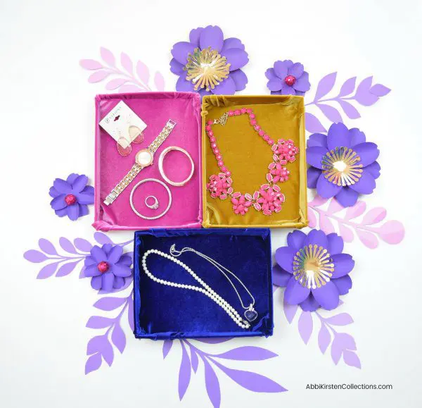 Cardboard storage boxes made into velvet drawer storage and jewelry organizers sit atop purple paper flowers against a white tabletop.