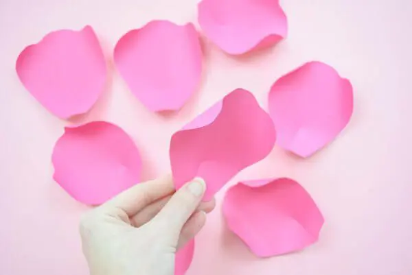 Small, curled pink paper petals lay on a tabletop ready for assembly. Abbi Kirsten's hands hold a finished small petal above the others.
