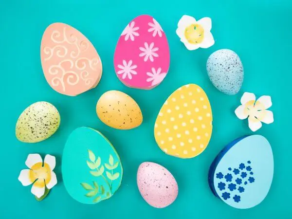 An overhead photo of red, orange, yellow, blue and more 3D paper Easter eggs with white flowers and real eggs as decorations on a teal background.