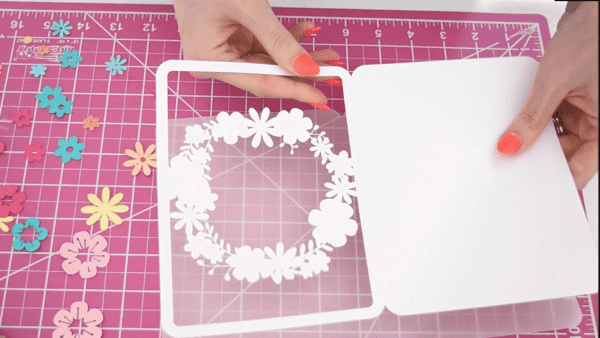 A white base for a DIY floral Mother's Day card alongside pastel flower cutouts on a pink workspace.