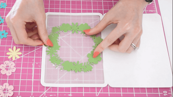 A green wreath being glued onto a white card atop a hot pink workspace.
