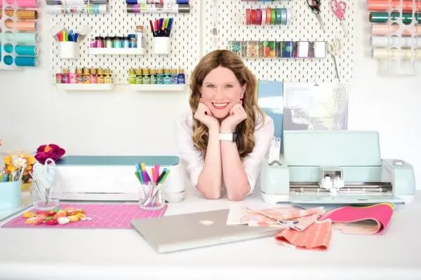 Abbi leans on her elbows, smiling in her craft room on a table of colorful supplies.