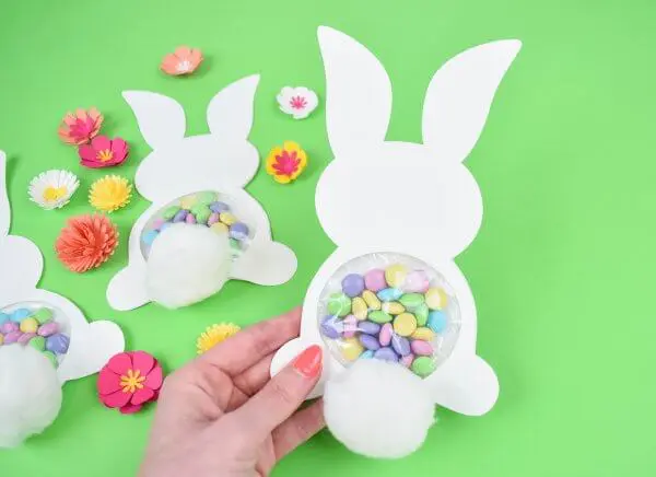 Abbi's hand holds one candy filled Easter Bunny craft. Two more, filled with pastel candy, lay on a light green backdrop surrounded by yellow, orange and white spring flowers. Free templates included!