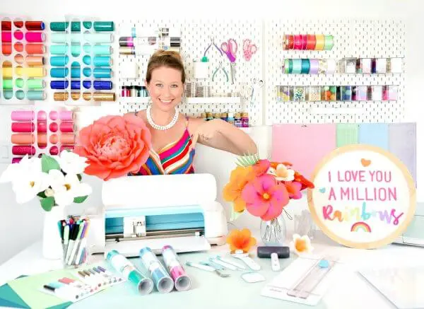 Abbi smiles in a rainbow sundress with a white pearl necklace. She stands in her craft room in front of a variety of organized craft supplies and behind her craft table area on which is a Cricut cutting machine, vinyl rolls, large and small orange and white paper flowers, and a sundry of other craft supplies. A circular craft says "I love you a million rainbows."