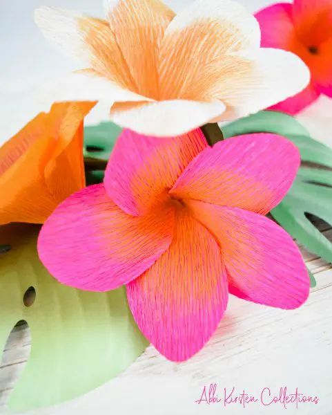 A Cricut machine was used to make these three small plumeria crepe paper flowers shown in a close-up with paper leaves on a light grey table. There is a pink flower with orange shading, a white flower with orange shading, and an orange flower facing away from the camera.