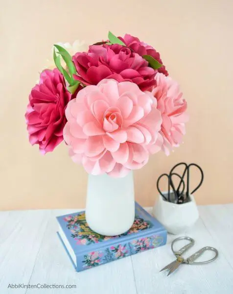 A white vase of paper Camellia roses in varying shades of pink with green paper leaves. The white vase is on a blue book, against a beige wall, and next to a white cup full of scissors.