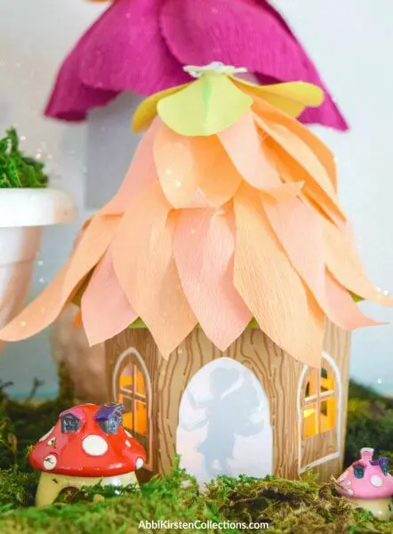 Behind painted mushrooms sits a paper fairy house with debossed paper walls and peach colored crepe paper flower petals for a roof. The silhouette of a fairy is seen in the doorway. 
