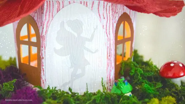 A closeup image of the door on a DIY fairy house, with a silhouette of a tiny, winged fairy dancing in the doorway. The house has paper windows, a crepe paper flower roof, and faux moss garden around the outside.