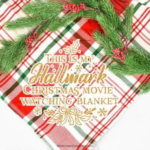 A red, white, and green plaid blanket with a white and gold iron-on vinyl decal that says "this is my Hallmark Christmas movie watching blanket" on it.