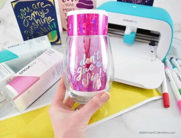 A personalize glass tumbler with hot pink smart vinyl that reads I don't give a sip. There is a blue Cricut Joy machine along with Cricut supplies in the background.