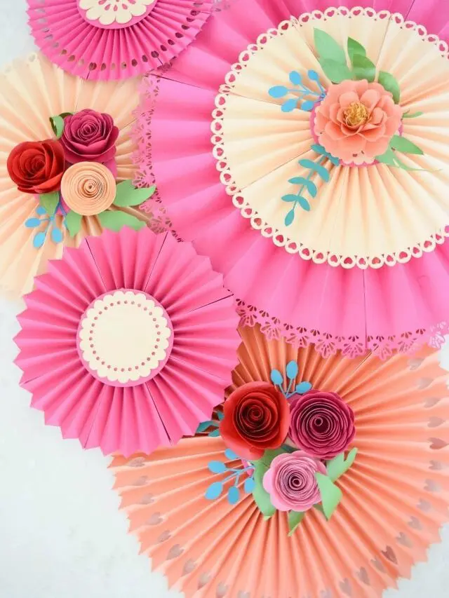 Pink and peach paper rosette fans hang overlapped on a white wall, with paper roses and greenery adorning the center of the decorations.