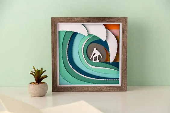 A Cricut Design Space craft project of a paper-layered surfer in waves of blue and green. The Cricut wavy edge blade was used to create this art piece. 