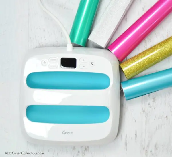 On top of the marble table lies a blue and white Easypress surrounded by green white, pink, gold and blue shimmering heat vinyl rolls. These hacks will help you create great looking Cricut vinyl crafts.