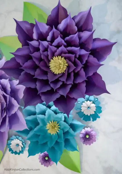 A close-up of a giant purple and blue amaryllis paper flowers, surrounded by smaller flowers and green leaves.