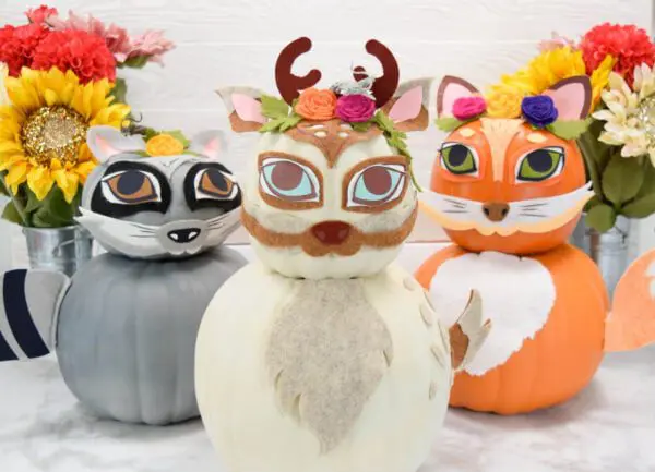 A deer made from two pumpkins sits in the foreground on a white floor. A gray raccoon and an orange fox sit in the back and to the side of the deer. Flowerpots sit int he background.