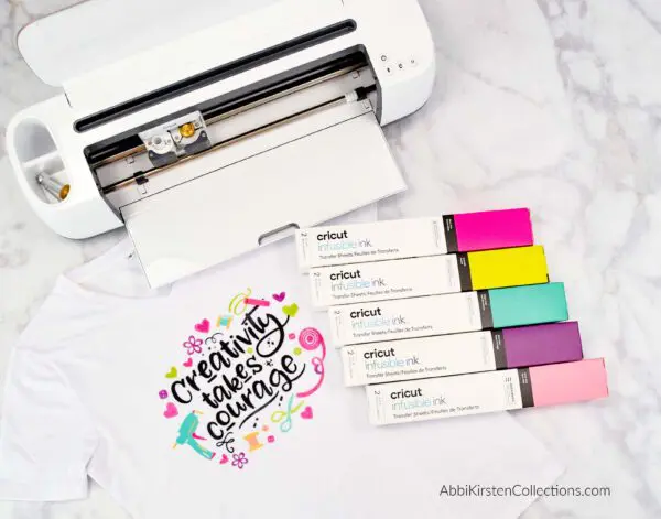 A photo of an open Cricut machine, cartons of infusible ink in multiple colors, and a white t-shirt with
"Creativity take courage" written in black text surrounded by colorful craft supply illustrations. Cricut Infusible Ink directions suggest using light-colored shirts for the best, most vibrant results.