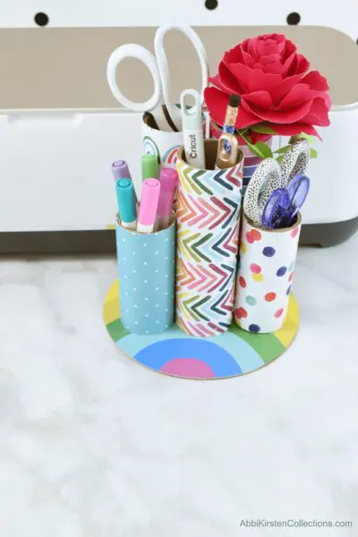 This pen organizer is made with upcycled cardboard tubes, each a different length, covered in washi tape and attached to a rainbow base. This desk organizer can hold pens, markers, scissors and more.
