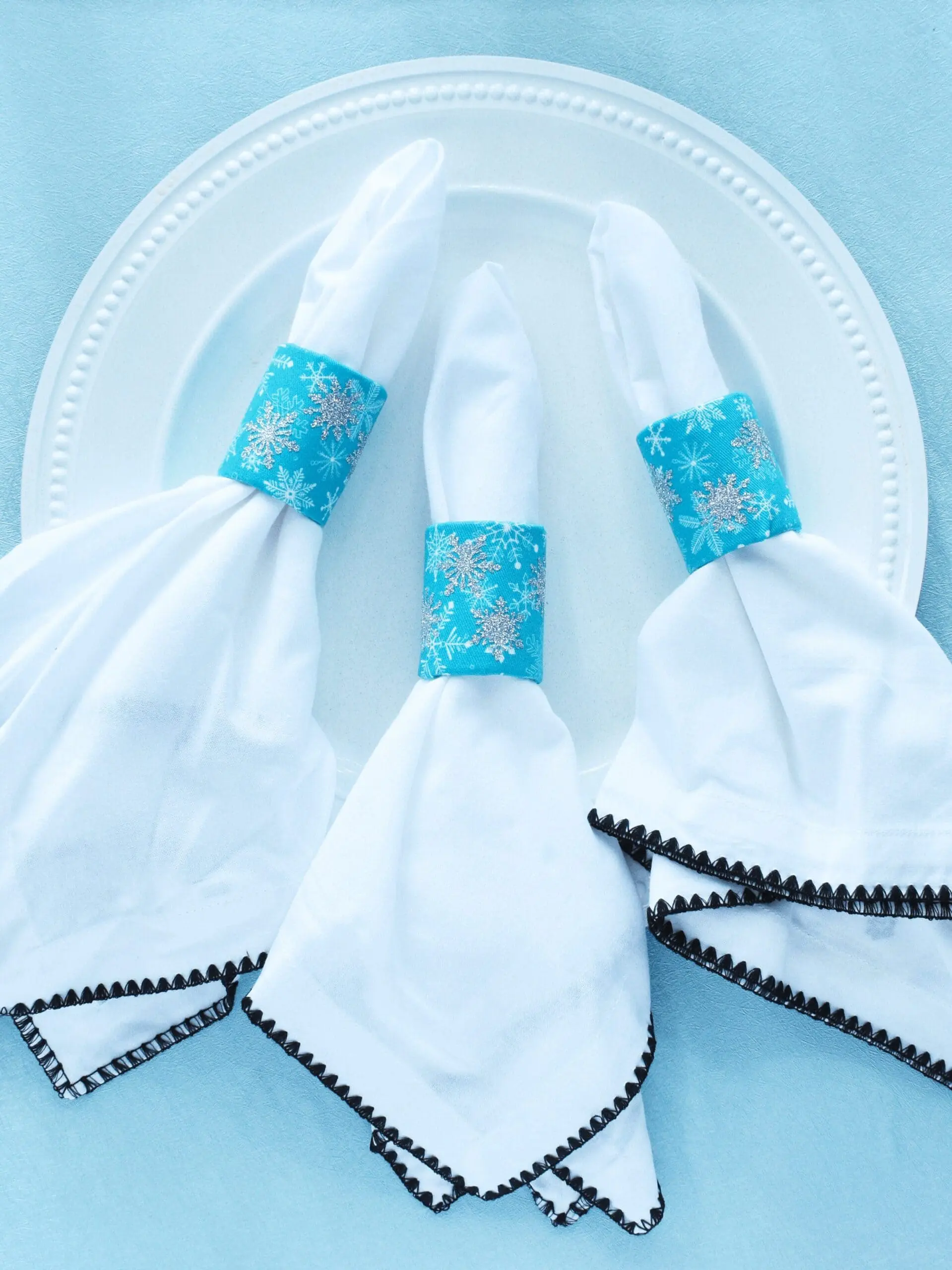 White napkins with black trim look festive and ready for a holiday tablescape thanks to upcycled cardboard tube napkin rings. The rings are covered in blue snowflake print fabric, and the napkins are against a white plate on a light blue background.