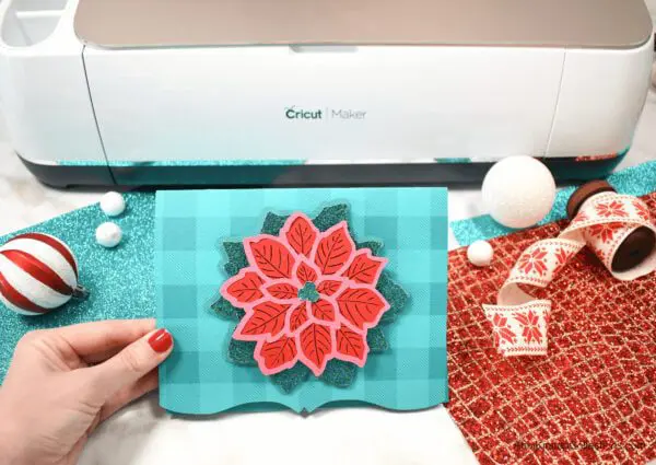 A gorgeous and festive Poinsettia Mandala Christmas Card held in front of a Cricut Maker cutting machine, surrounded by glitter craft paper and Christmas decorations.
