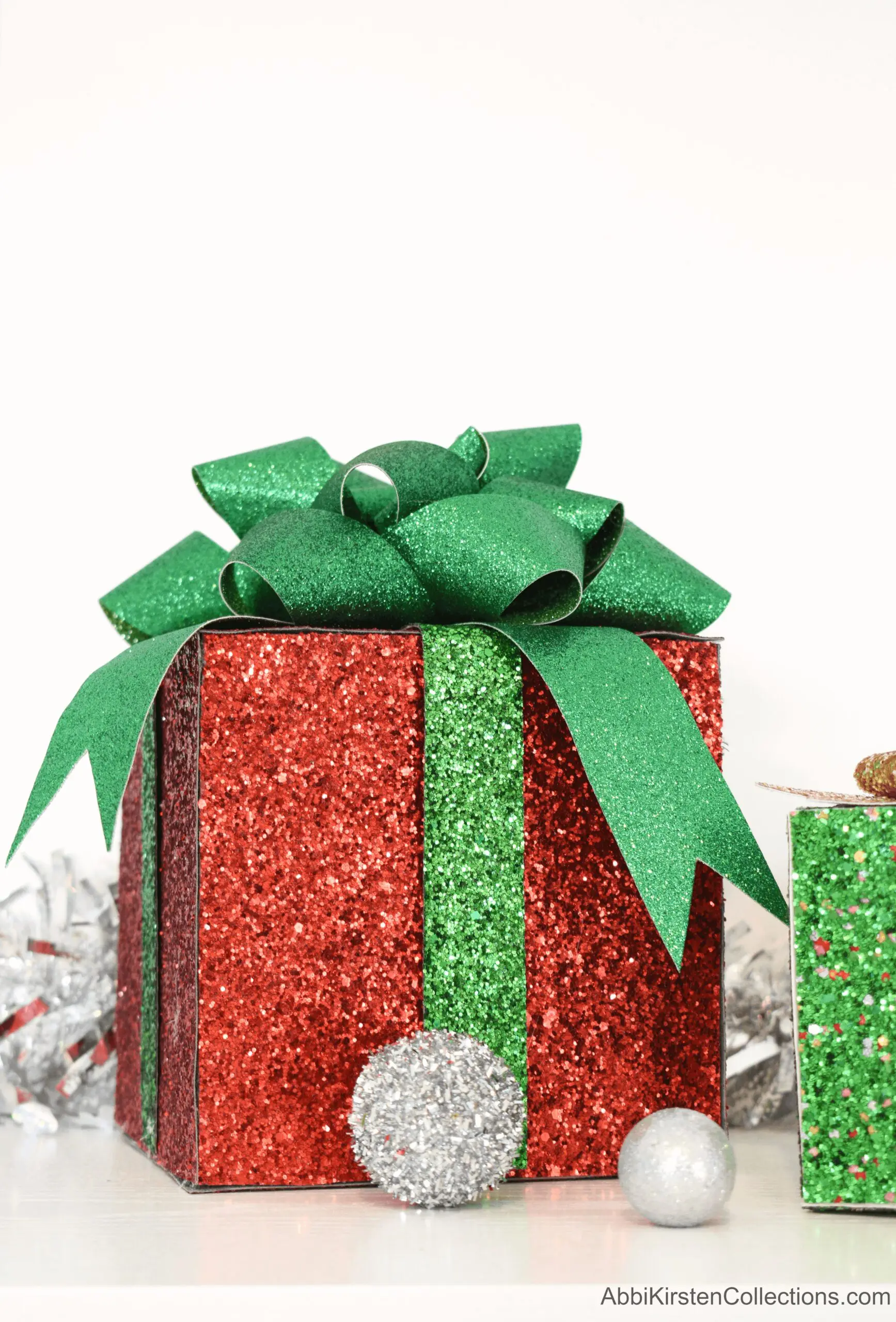 A glittery red decorative gift box with a green bow sits on the floor surrounded by silver tinsel.