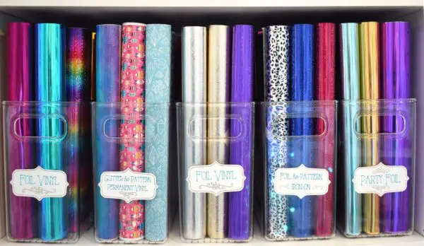 A closeup of foil transfer labels on clear bins lined up on a bookcase full of various colors of vinyl rolls. Labelling and organizing helps crafting take less time.