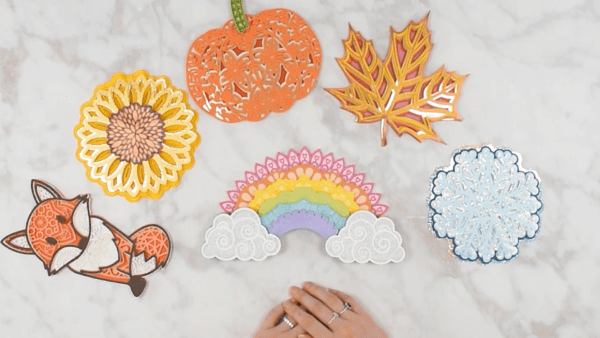 The finished 3D rainbow mandala, laid on a table, surrounded by 3D mandalas in the shapes of a fox, pumpkin, sunflower, leaf and snowflake.