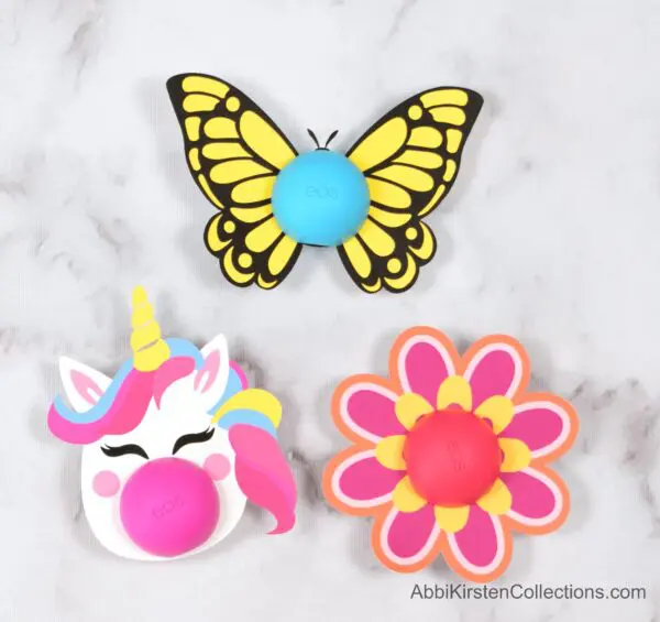A close-up overhead view of the three free printables for Eos lip balms include a unicorn, a pink flower, and a yellow butterfly.