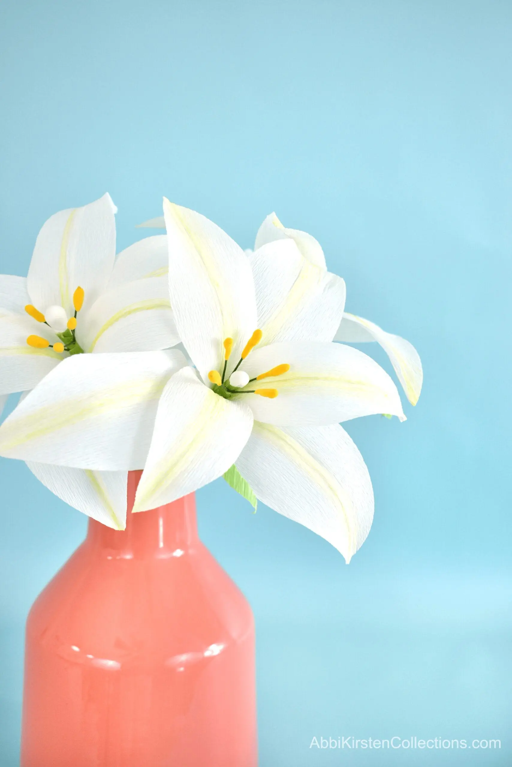 A bundle of crepe paper Easter lily flowers in a opaque salmon-colored ceramic vase in front of a light blue paper backdrop.