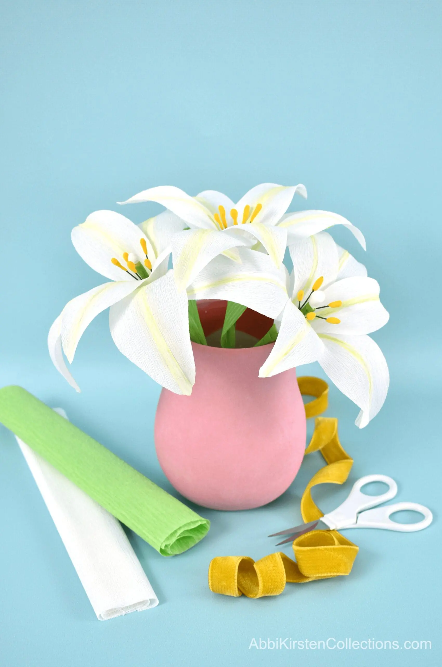A finished bouquet of three white crepe paper lilies with green leaves in a pink vase. Rolls of white and green crepe paper, detail scissors and yellow ribbon lay in front of the vase on a light blus paper backdrop.