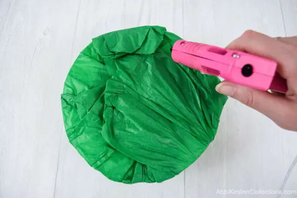 A foam bouquet base wrapped in green tissue paper. A woman’s hand is hot gluing the edges of the tissue paper together so it stays secure.
