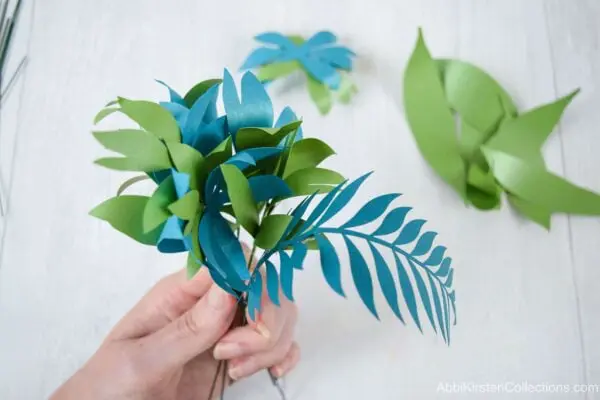 A bundle of greenery in various shades of green is held in a woman’s hand. This greenery will be placed among the paper flowers in the arrangement or bouquet.