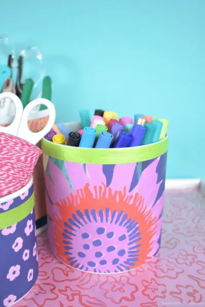 A cylinder vase filled with markers and decorated with pink, red and purple wrapping paper and green ribbon.