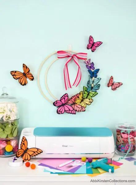 A paper butterfly wreath hangs on a light blue wall, surrounded by colorful paper butterflies fluttering around. Cricut supplies and cardstock are laid out on a table below.