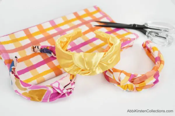 Three multicolored satin fabric headbands are displayed on a checkered piece of fabric. With this tutorial you can make a variety of no-sew top knot headbands.