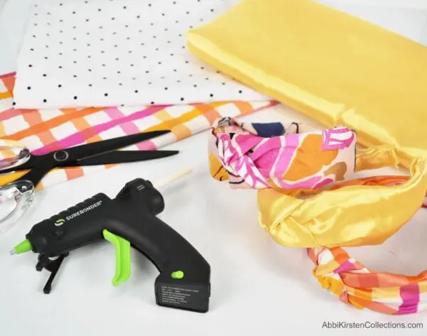 A tabletop display of multiple headbands and the varying colors of fabrics used to make them, as well a pair of fabric scissors and Surebonder Mini glue gun.