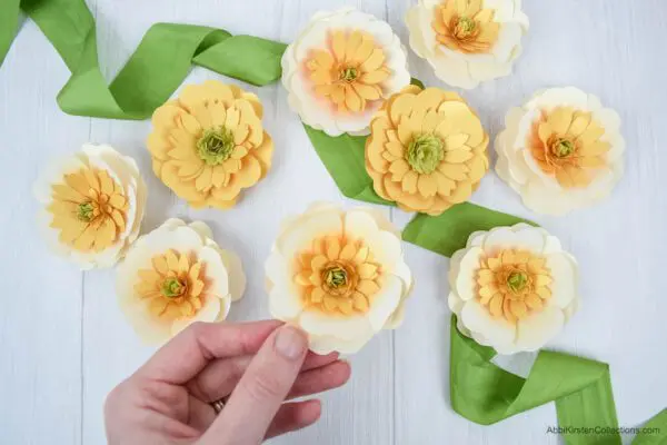 A bright green ribbon underneath nine pale yellow buttercup paper flowers with light green centers displayed on a white table.