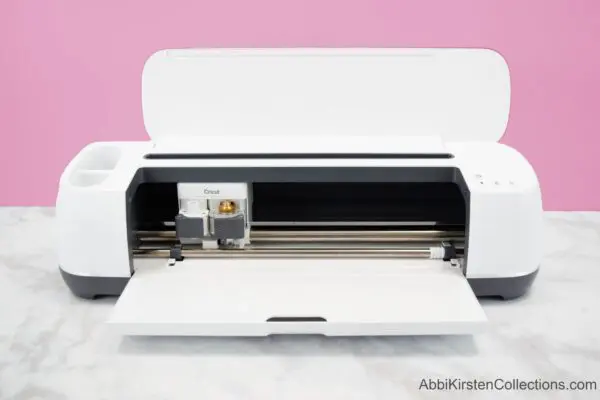 A white Cricut Explorer machine on a clean marble tabletop and a dusty rose background. This machine is easy to learn.