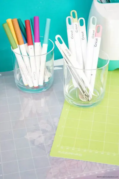 Cricut pens and other accessories you will need to use your Cricut machine. The pens are in clear glass cups and rest on top of Cricut mats.