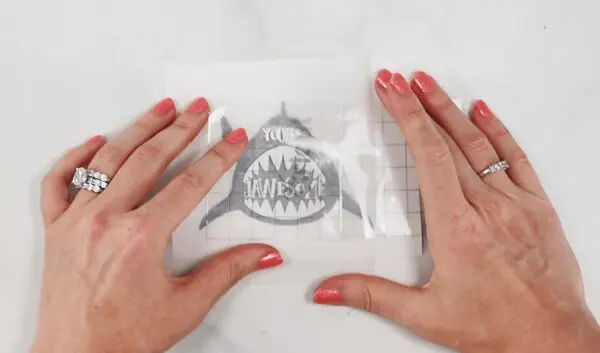 A woman's hands hold transfer tape over a vinyl sticker of a shark's open mouth. Parchment paper is just one tip to making Cricut vinyl crafts easier.