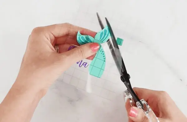 A woman's hands use scissors to trim down the transfer tape on a vinyl mermaid's tail. Burnishing is a helpful hack for Cricut vinyl crafts.