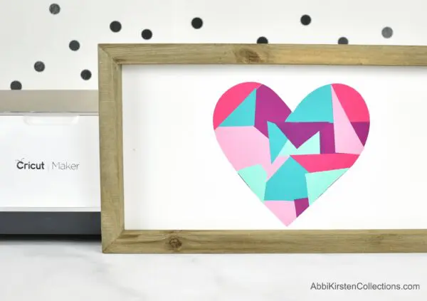 A wooden frame with a white background and red, teal, pink and purple colored heart in the center. One Cricut vinyl hack is to create art using your vinyl scraps.