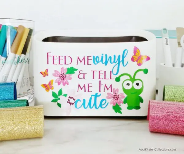 A closeup of a cute mini vinyl scraps trashcan. It has a frog, flowers, and butterflies on the white side with the words "Feed me vinyl and tell me I'm cute." Just one vinyl hack of 25 to use with your Cricut machine.