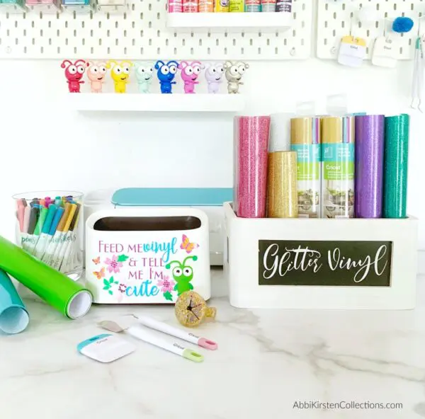 A closeup of a tabletop hosting several craft supplies like the vinyl scraps trashcan, markers and a wooden bin storing glitter vinyl rolls. Storing and organizing vinyl rolls is one tip to make vinyl and Cricut crafting easier.
