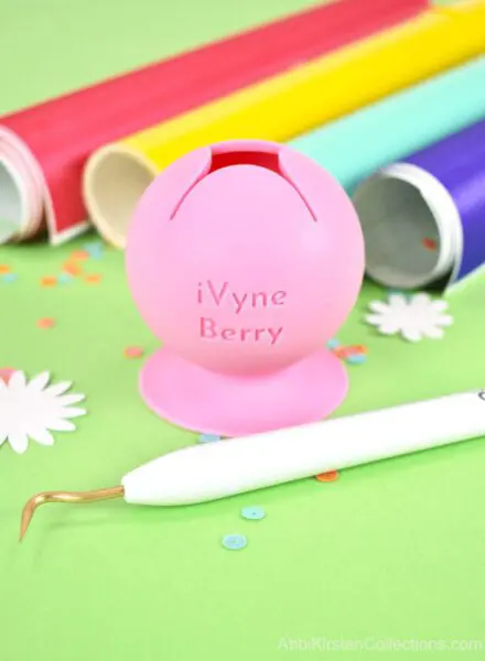 Tips for weeding Cricut vinyl include using this iVyne Berry Tool. A closeup of the pink berry tool, a standard weeding tool an primary color vinyl rolls on a green table top.