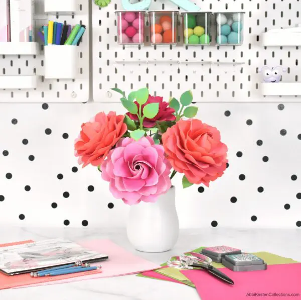 Paper stemmed roses in shades of red and pink are arranged in a white vase. This full garden bloom paper rose tutorial can be used to make any color of rose you want for any occasion.