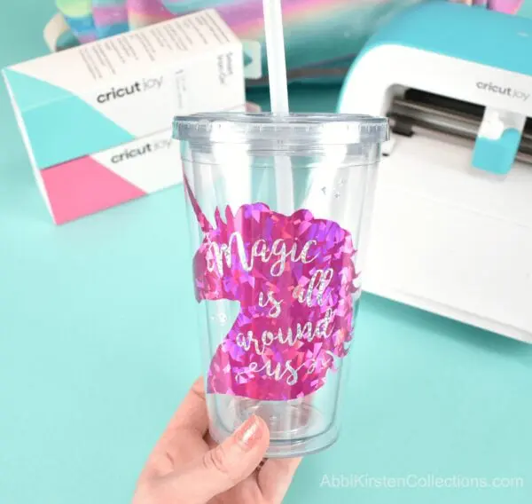 A clear plastic drink tumblr with a holographic pink unicorn vinyl decal that says "magic is all around us"