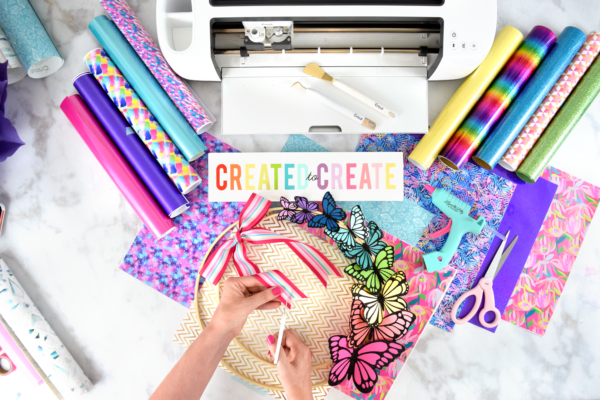A bird's eye view of Abbi Kirsten as she decorates a hat with paper butterflies and ribbons, with rolls of vinyl and a Cricut machine in front of her. 