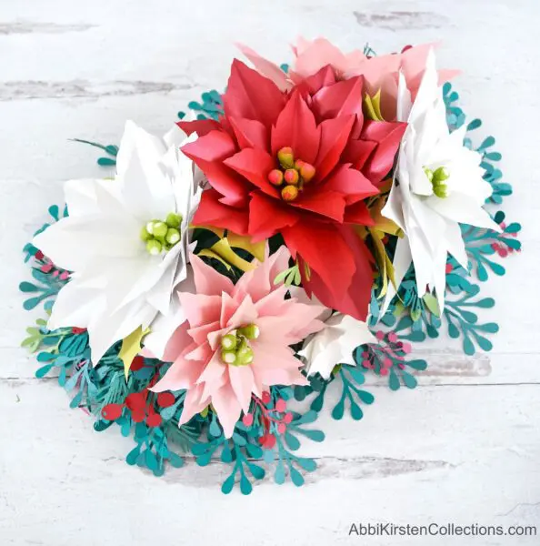 A colorful paper poinsettia flower arrangement made with white, pink, and red poinsettia paper flowers and blue and red leaves.