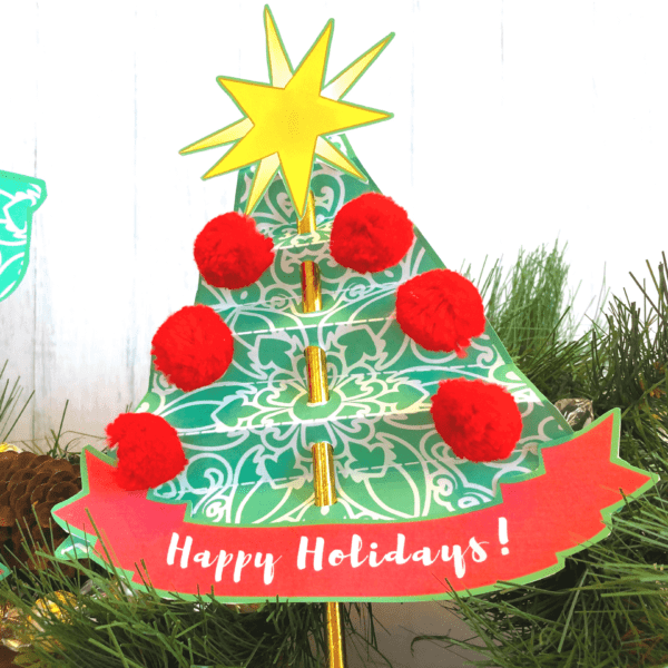 An accordion-folded green and white paper Christmas tree is adorned with pom-poms, a “Happy Holidays” banner, and a bright yellow paper star. This simple craft is great for kids.