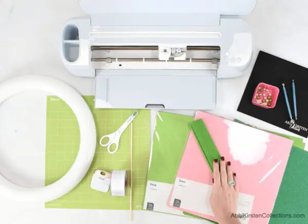 In front of a pink wall, on a white table, sit colorful Christmas trees, a string of white baubles, and small wrapped presents. A paper poinsettia and holly wreath hangs on the wall.